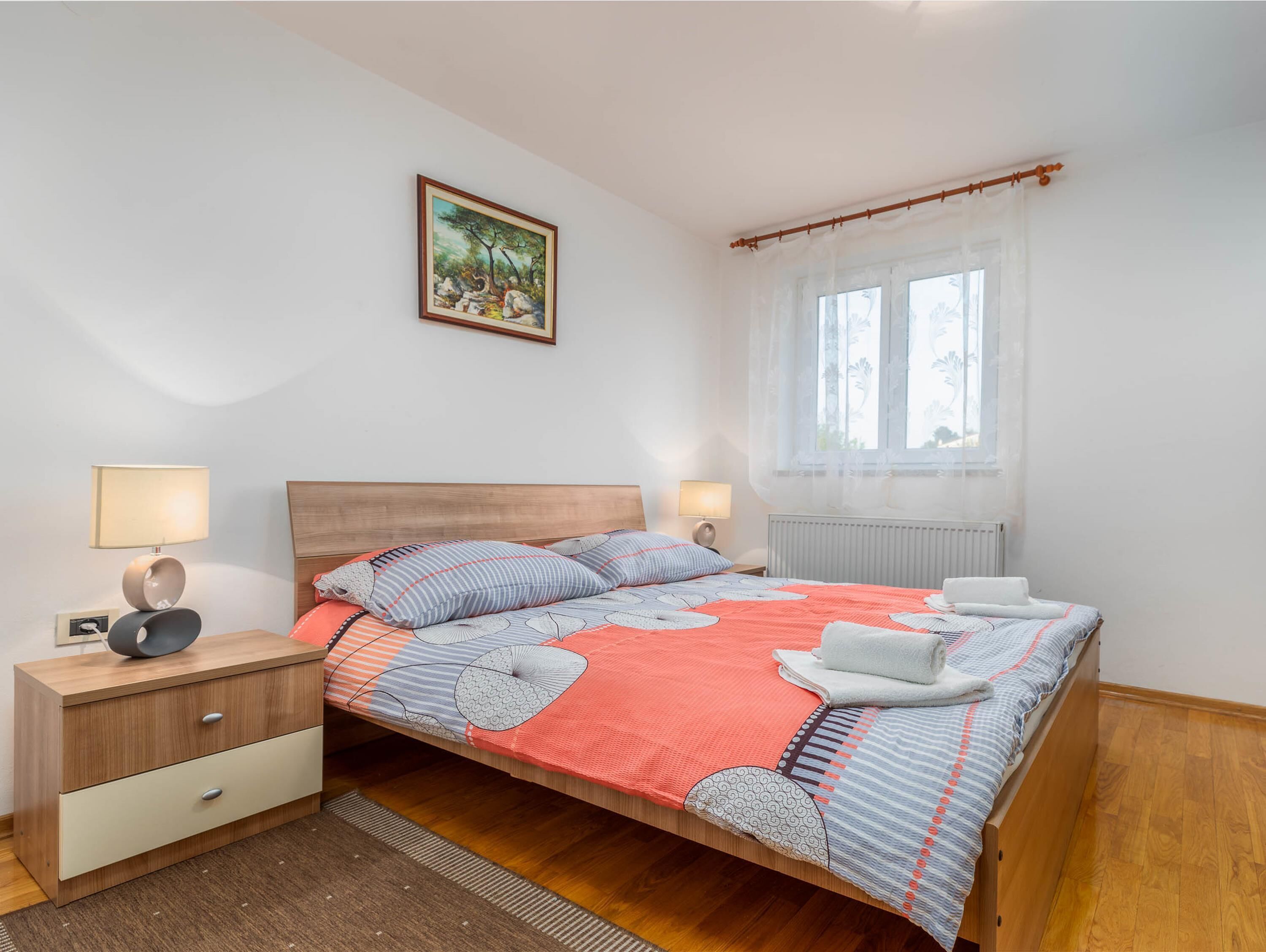 Un dormitorio ordenado con cama, mesitas de noche, una ventana con cortinas y un cuadro en la pared.