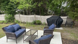 Eine Terrasse mit Korbmöbeln, einem Glastisch und einer überdachten Grillgegend.
