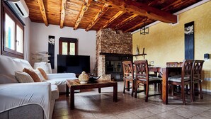 Un salon chaleureux avec une cheminée en pierre, des poutres apparentes en bois au plafond, un canapé, un téléviseur et un coin repas avec des chaises.