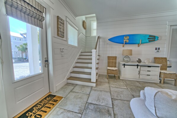 A staircase leading to a second floor, a white door with a doormat, a blue surfboard on the wall, and a white sofa in the living area.