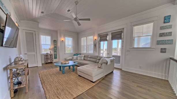 A spacious living room with a beige sectional sofa, a round coffee table, and a woven rug. There are multiple windows with blinds, a ceiling fan, and a television mounted on the wall. - Hangtime Inlet Beach/Next to Rosemary Beach (Inlet Beach)