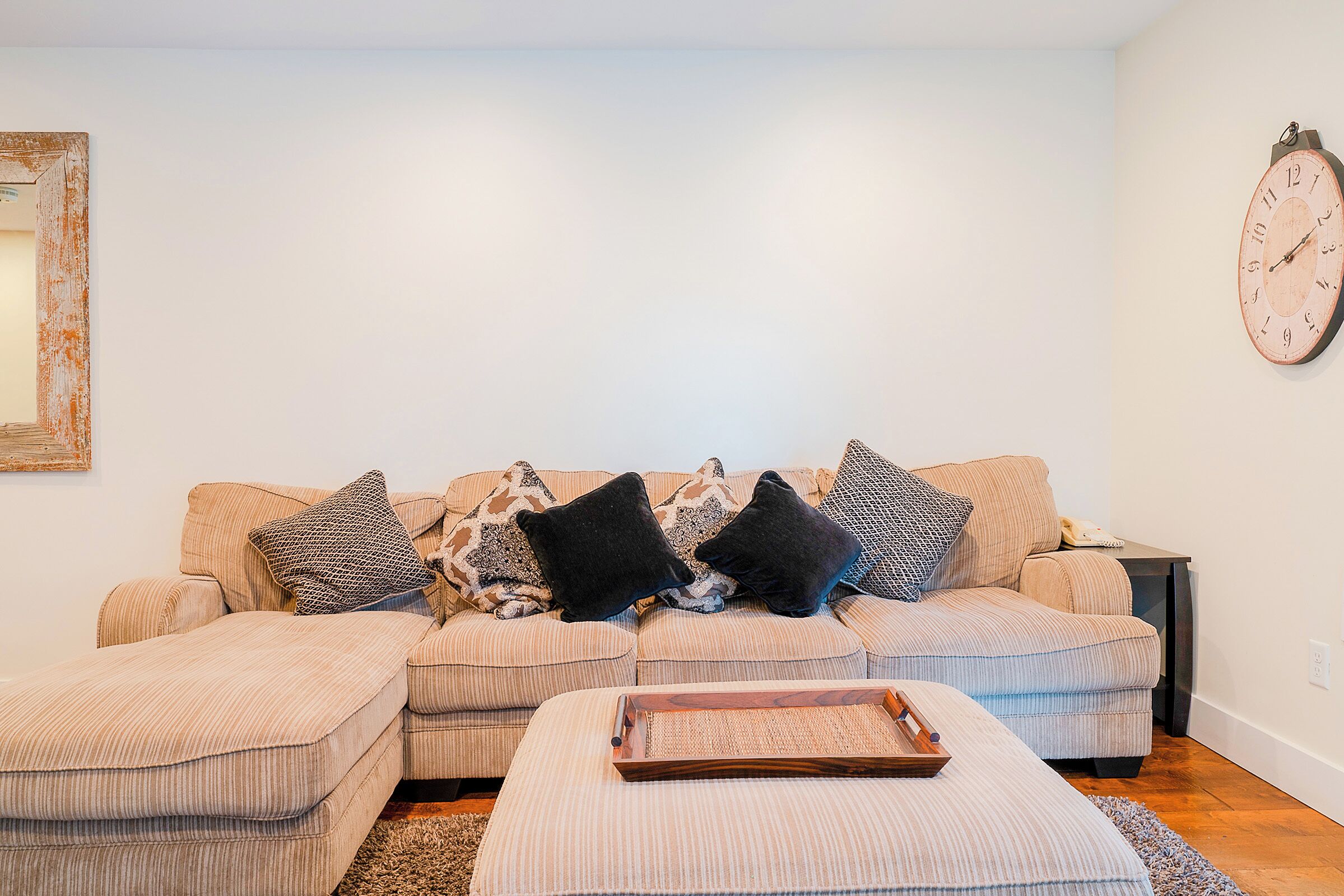 A living room with a beige sofa, two ottomans, a wooden tray, and a clock on the wall.
