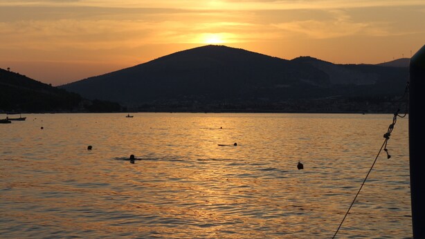 Un coucher de soleil sur une étendue d’eau calme, avec des bateaux et une chaîne de montagnes en arrière-plan.