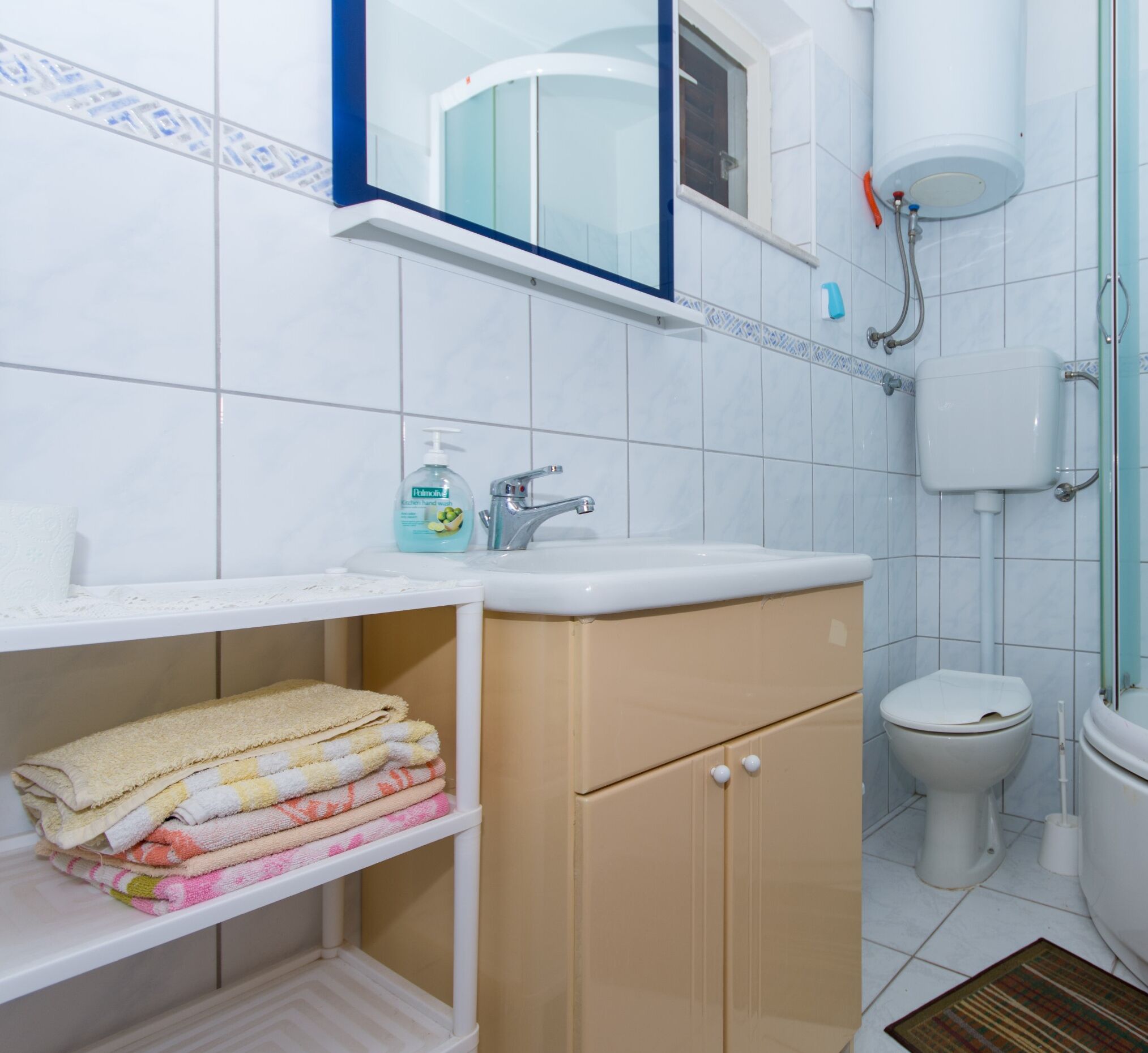 A bathroom with a sink, toilet, and shower. There is a shelf with towels and a bottle of hand soap.