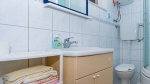 Une salle de bain équipée d’un lavabo, de toilettes et d’une douche. On y trouve une étagère avec des serviettes et un flacon de savon pour les mains.