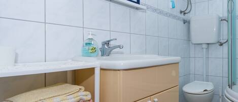 A bathroom with a sink, toilet, and shower. There is a shelf with towels and a bottle of hand soap.
