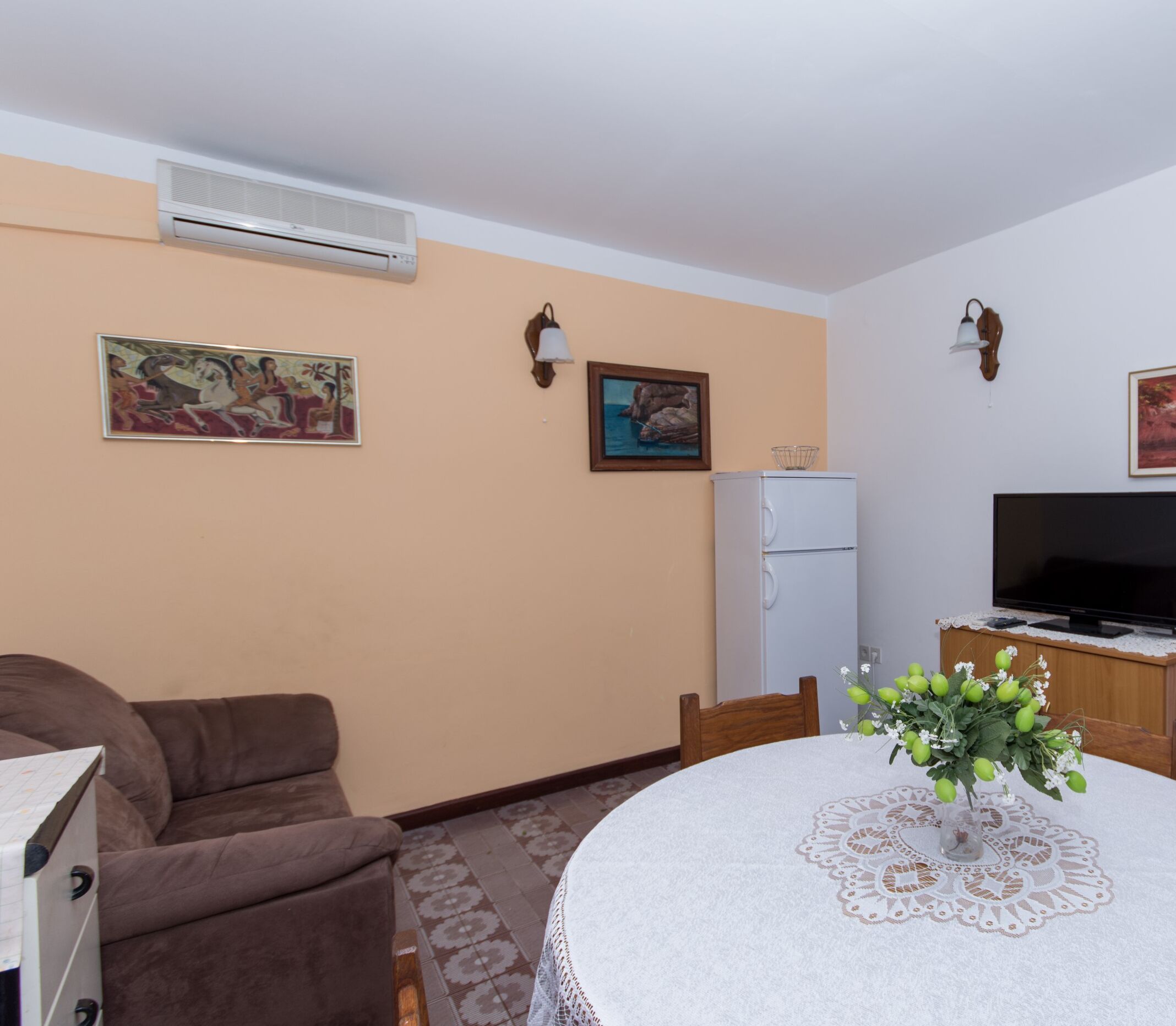 A living room with a brown sofa, a dining table with a lace tablecloth, a TV on a wooden cabinet, and a refrigerator.