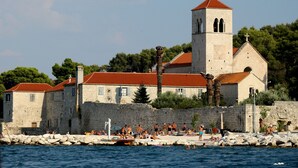 Un bâtiment historique en pierre, doté d’un toit recouvert de tuiles rouges, d’un clocher et situé en bord de mer, où l’on peut voir des gens nager et prendre des bains de soleil.