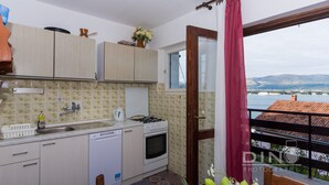 A compact kitchen with a sink, stove, and dishwasher, a wooden dining table with chairs, and a view of the sea through a balcony with a red curtain. - A2 Large apt With the big Terrace & Great sea View (Trogir)
