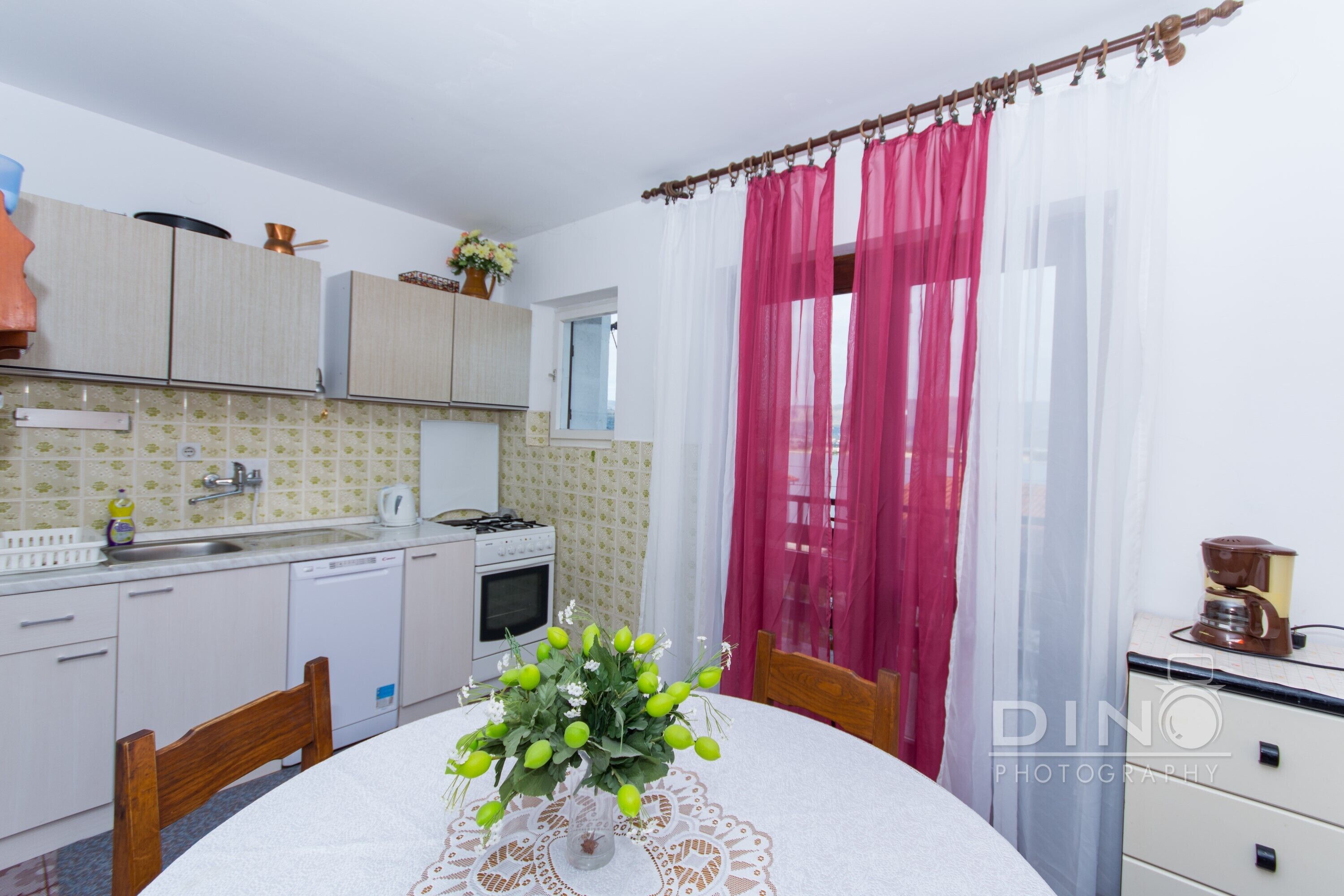 A kitchen with white cabinets, a stove, and a sink. There is a table with a vase of flowers.