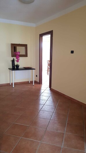 Una habitación amplia con suelo de baldosas, una mesita con una planta en maceta, un espejo y una puerta que da a otra zona.