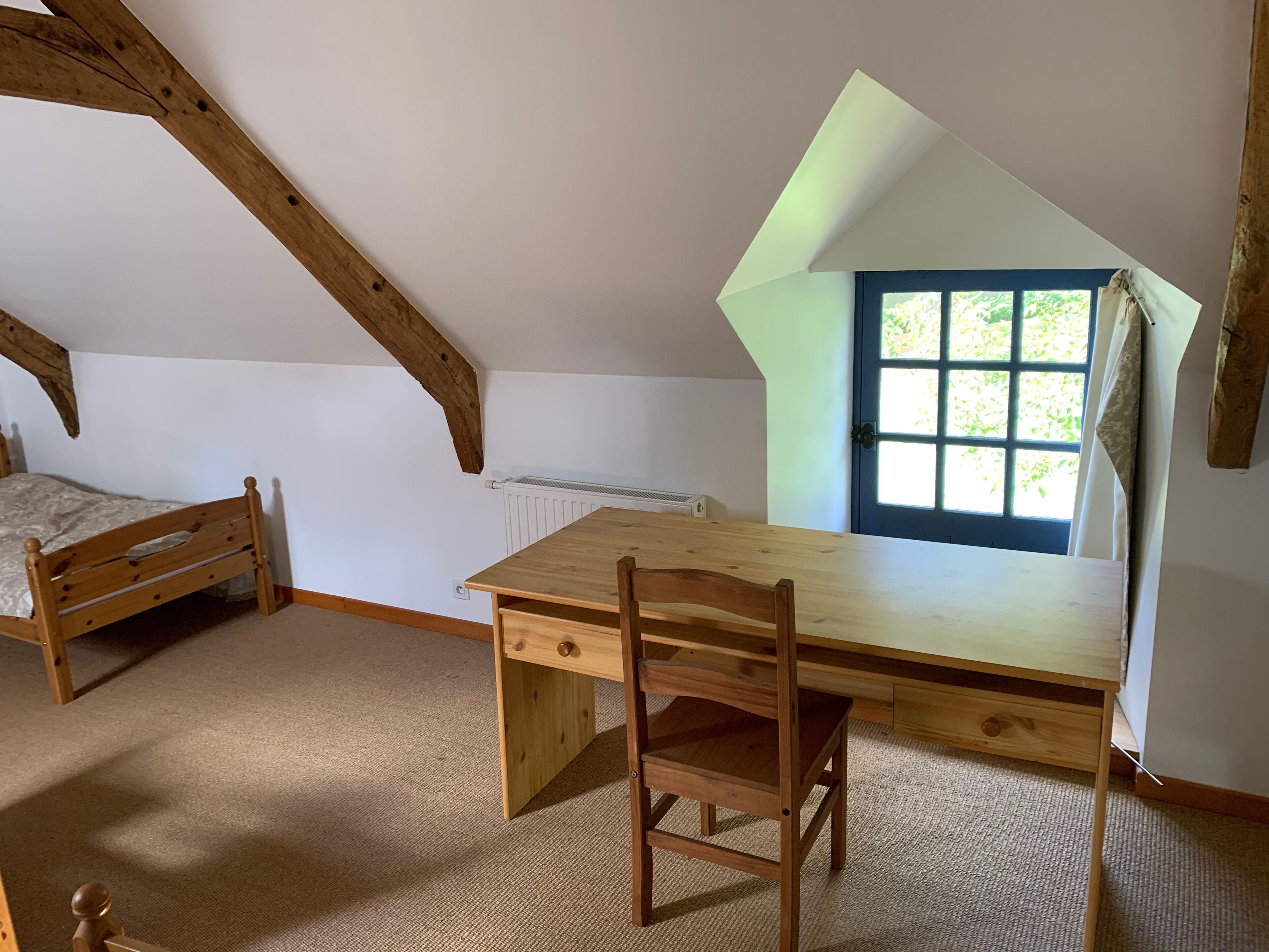 A room with a sloped ceiling, a wooden desk with a chair, a bed, and a window with a view of greenery.