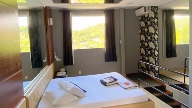 A hotel room with a bed, a small table, a window with curtains, and a view of greenery outside. - Cequisabe Motel (Juiz de Fora)