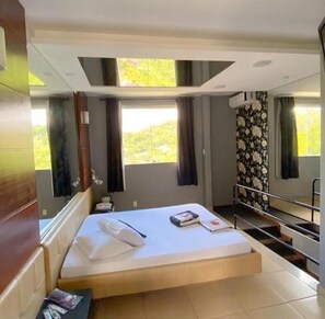 A hotel room with a bed, a small table, a window with curtains, and a view of greenery outside. - Cequisabe Motel (Juiz de Fora)
