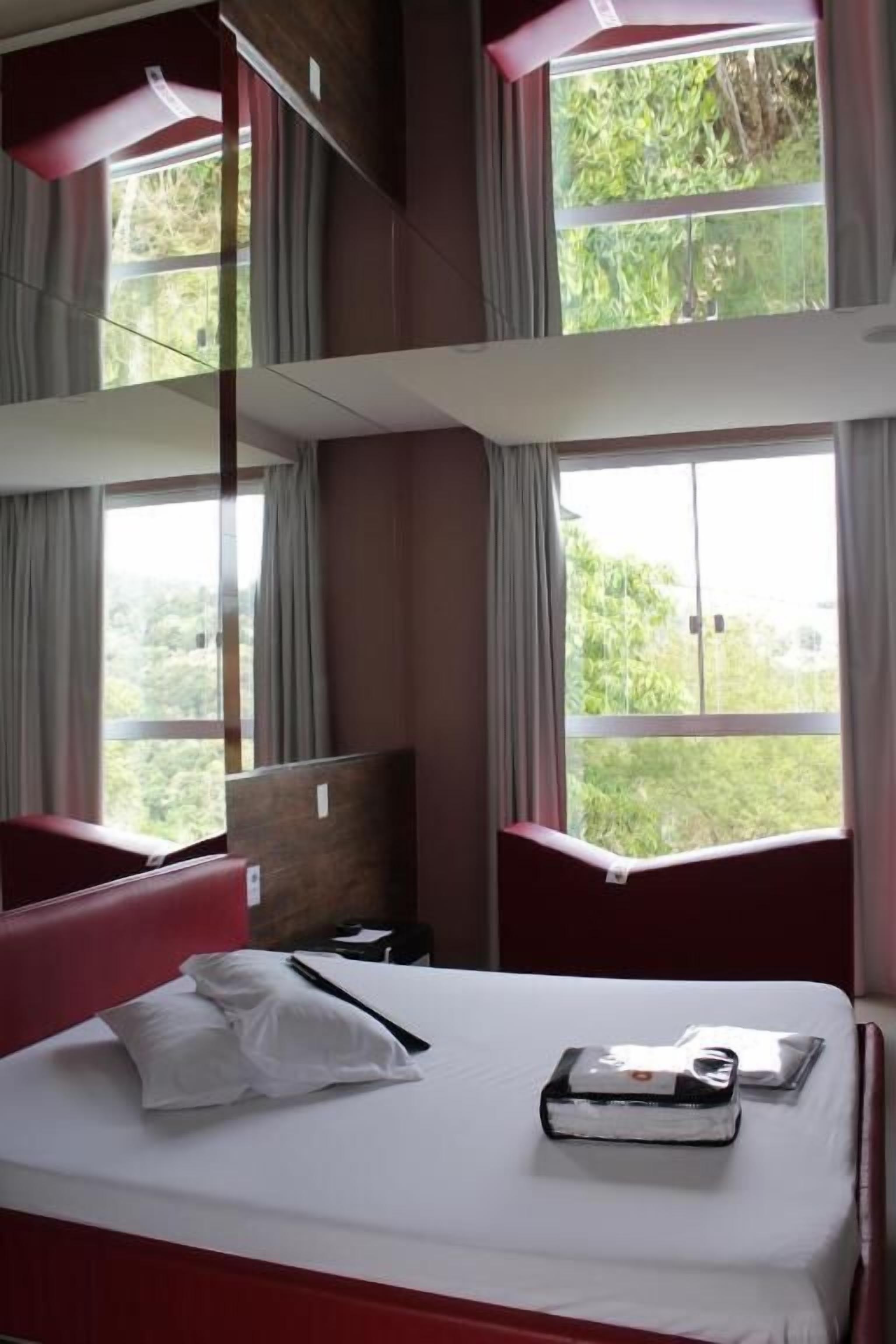 Quarto de hotel com cama, uma cadeira vermelha e janelas amplas com vista para o verde lá fora.