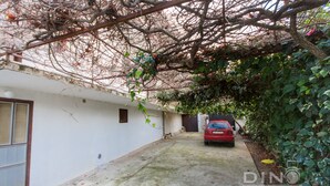 A narrow alleyway with a red car parked on the right, surrounded by greenery and a white building with a window and a door. - A1 Large apt With the big Terrace & Great sea View (Trogir)