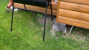 A black barbecue grill with a metal stand, positioned on a grassy lawn next to a wooden fence and a wooden structure. - Chata pri Jazere 1 - Lake Side Cottage (Horná Potôn)