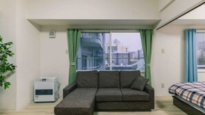 A room with a sectional sofa, a bed with a plaid bedspread, a window with green curtains, and an air conditioning unit. - IKminami6jo residence 601 (Sapporo)