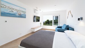 Una camera da letto moderna con un letto grande, un divano blu, un quadro che raffigura una scena costiera e una finestra vista sul verde.