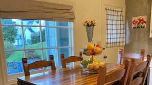 Un coin repas avec une table et des chaises en bois, une fenêtre donnant sur des maisons et un panier de fruits posé sur la table.