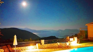 A pool area with stone flooring, a stone wall, and a metal fence. There are lounge chairs and an umbrella. - Executive Villa Scorpidi With Private Pool (Lefkada)