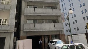 Un edificio de varios pisos con una entrada central, un guardia de seguridad y un coche blanco estacionado al frente.