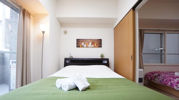 Una cama bien hecha con cubrecama verde y sábanas blancas, una mesita de noche pequeña con una lámpara y una ventana con cortinas.