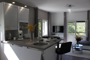 Een moderne keuken met een inbouwoven, een koffiezetapparaat en een spoelbak. Er is een eethoek met een tafel en stoelen, en een televisie aan de muur bevestigd.