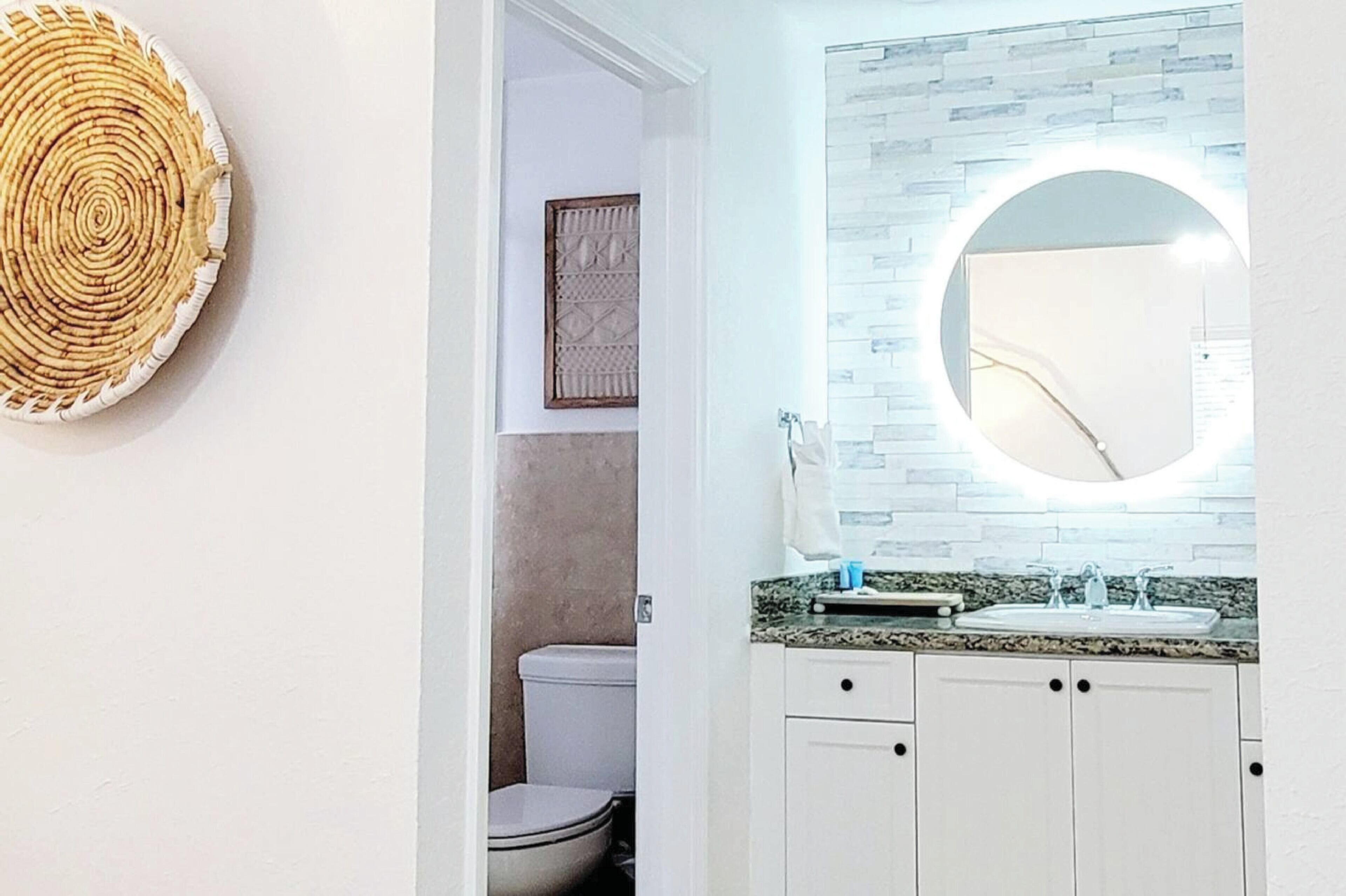 A bathroom with a white toilet, a sink with a granite countertop, and a round mirror with a light.