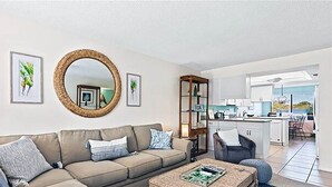 A modern living room with a sofa, a coffee table, and a large mirror. There is a kitchen in the background with white cabinets and a dining area with a table and chairs. - Sandpebble 2B (Sanibel)