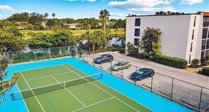 A tennis court with a net, surrounded by a fence, with a parking area and buildings in the background. - Sandpebble 2B (Sanibel)