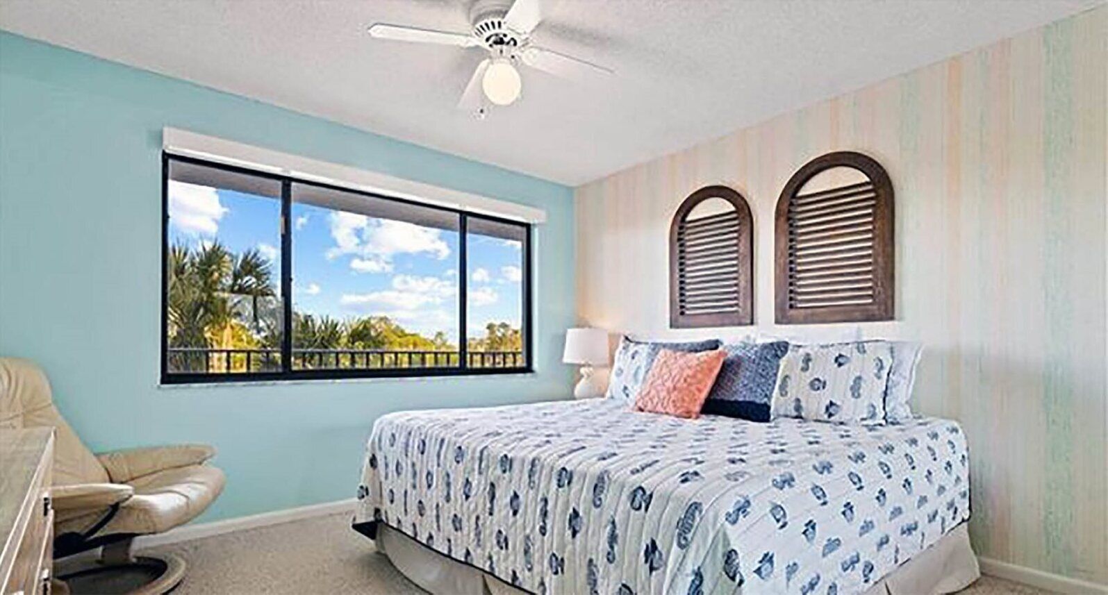 Un dormitorio con un ventanal grande, una cama con ropa de cama estampada, un ventilador de techo y vistas a palmeras.