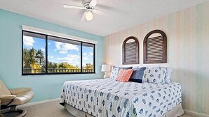 A bedroom with a large window, a bed with patterned bedding, a ceiling fan, and a view of palm trees. - Sandpebble 2B (Sanibel)