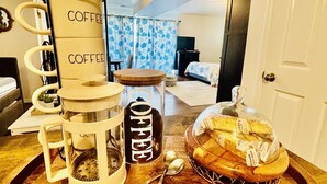Una zona de cocina con cafetera, cuchara, frasco de vidrio y tabla de madera con hogaza de pan.