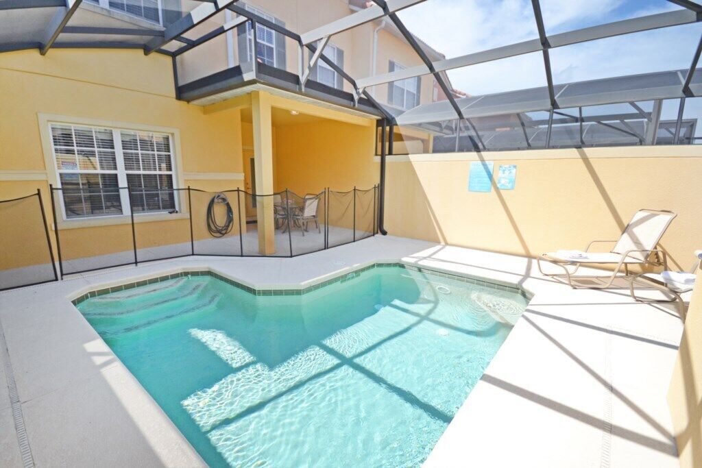 A pool area with a clear blue pool, a covered seating area with chairs, and a yellow wall.