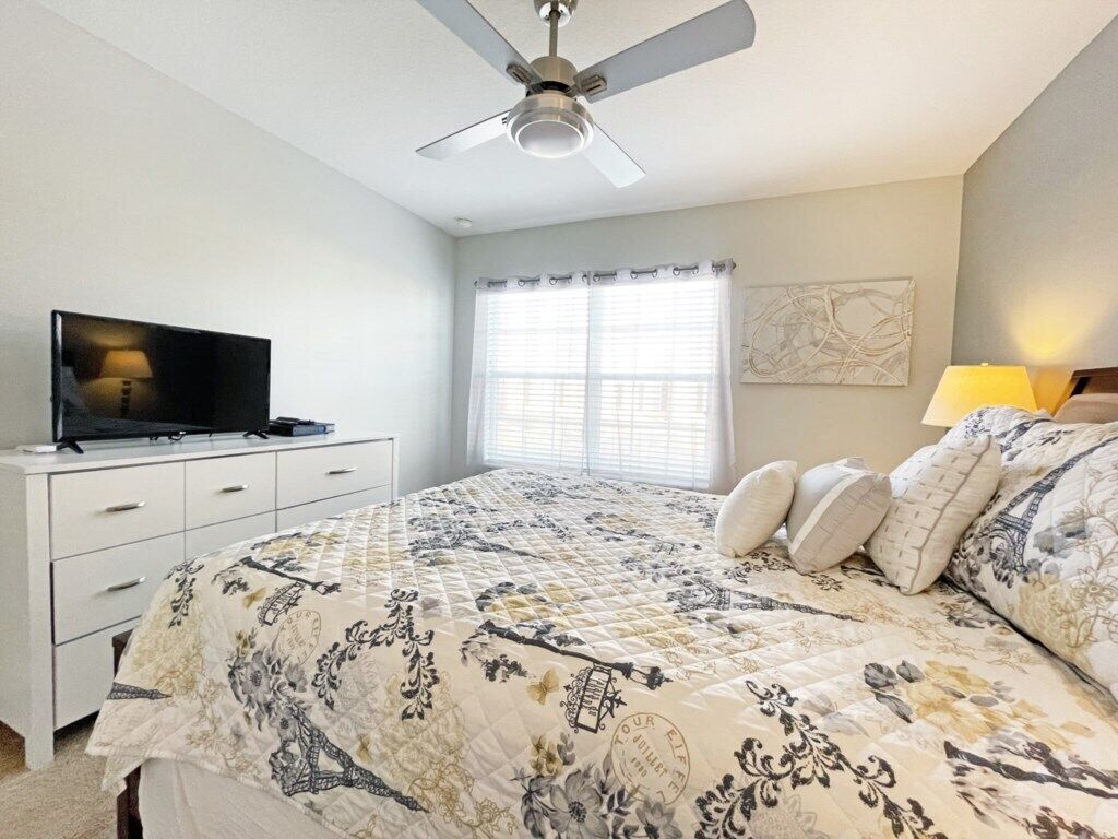 A bedroom with a bed, a ceiling fan, a television on a dresser, and a window with blinds.