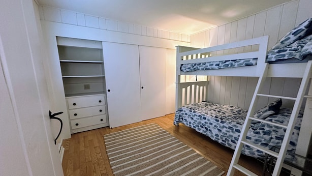 A bunk bed with a patterned bedspread, a wooden floor, a striped rug, and a white wardrobe with multiple drawers.