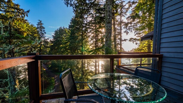 A glass table and chairs on a balcony with a view of a forest and a house.