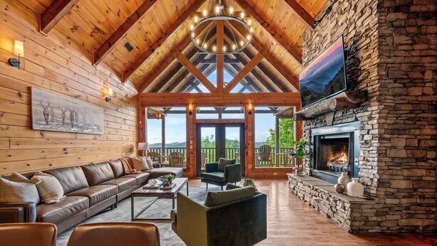 A spacious living room with a stone fireplace, wooden ceiling beams, and a large window overlooking a scenic landscape. - Massive Views+Indoor Pool+Movie Theater+Fire Pit (Sevierville)