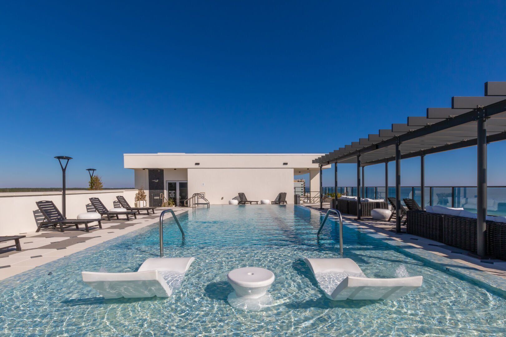 A rooftop infinity pool with lounge chairs, a covered seating area, and a modern building in the background.