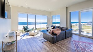 En rummelig stue med en stor grå sofa, et lille rundt bord og en grøn lænestol. Store vinduer giver udsigt over stranden og havet.