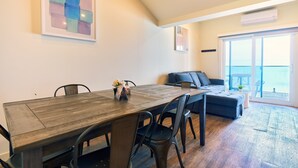 A modern living room with a wooden dining table, dark chairs, a sofa, and a large window offering a view of the sea.
