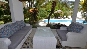 Ein überdachter Außenbereich mit zwei grauen Sofas und einem Couchtisch in der Mitte, mit Blick auf einen Pool und üppiges Grün.