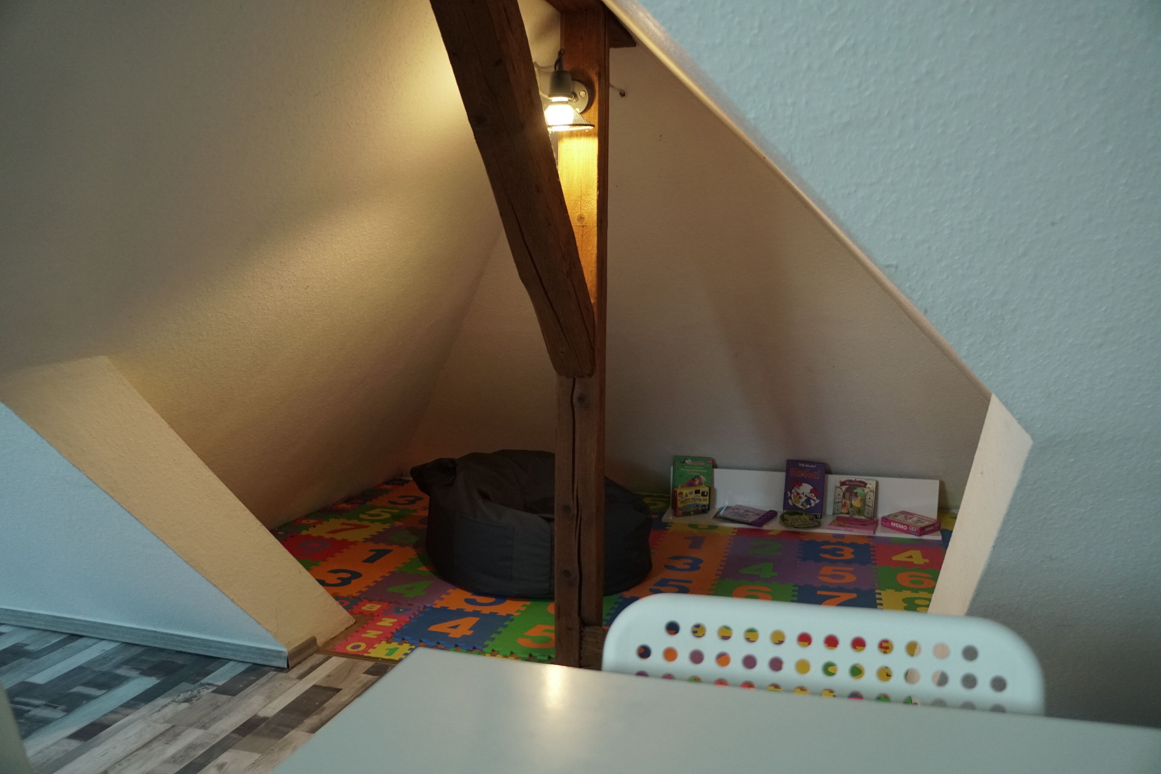 Ein kleines Dachzimmer mit schrägen Wänden, einem Sitzsack, einer bunten Spielmatte und einem weißen Stuhl mit Punktmuster.