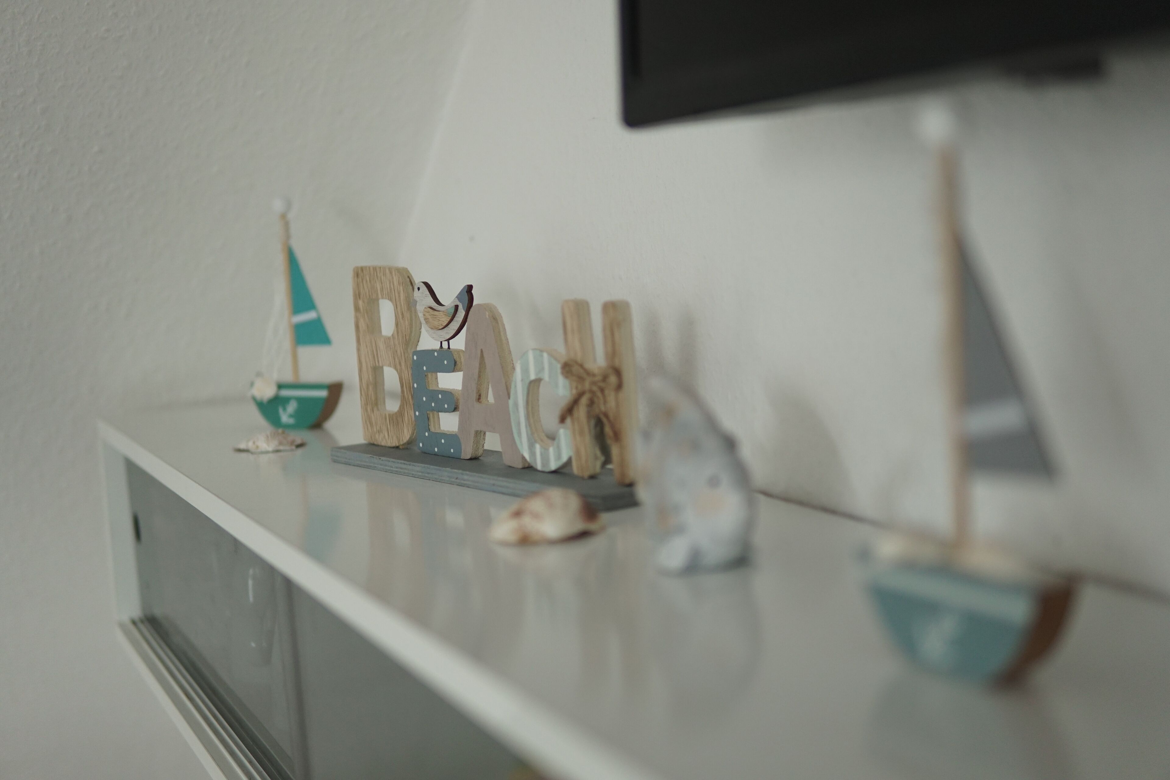 Eine weiße Kommode mit Dekorationsobjekten, darunter Holzbuchstaben, die ‚BEACH‘ buchstabieren, ein kleines Segelboot und ein Schiffmodell.