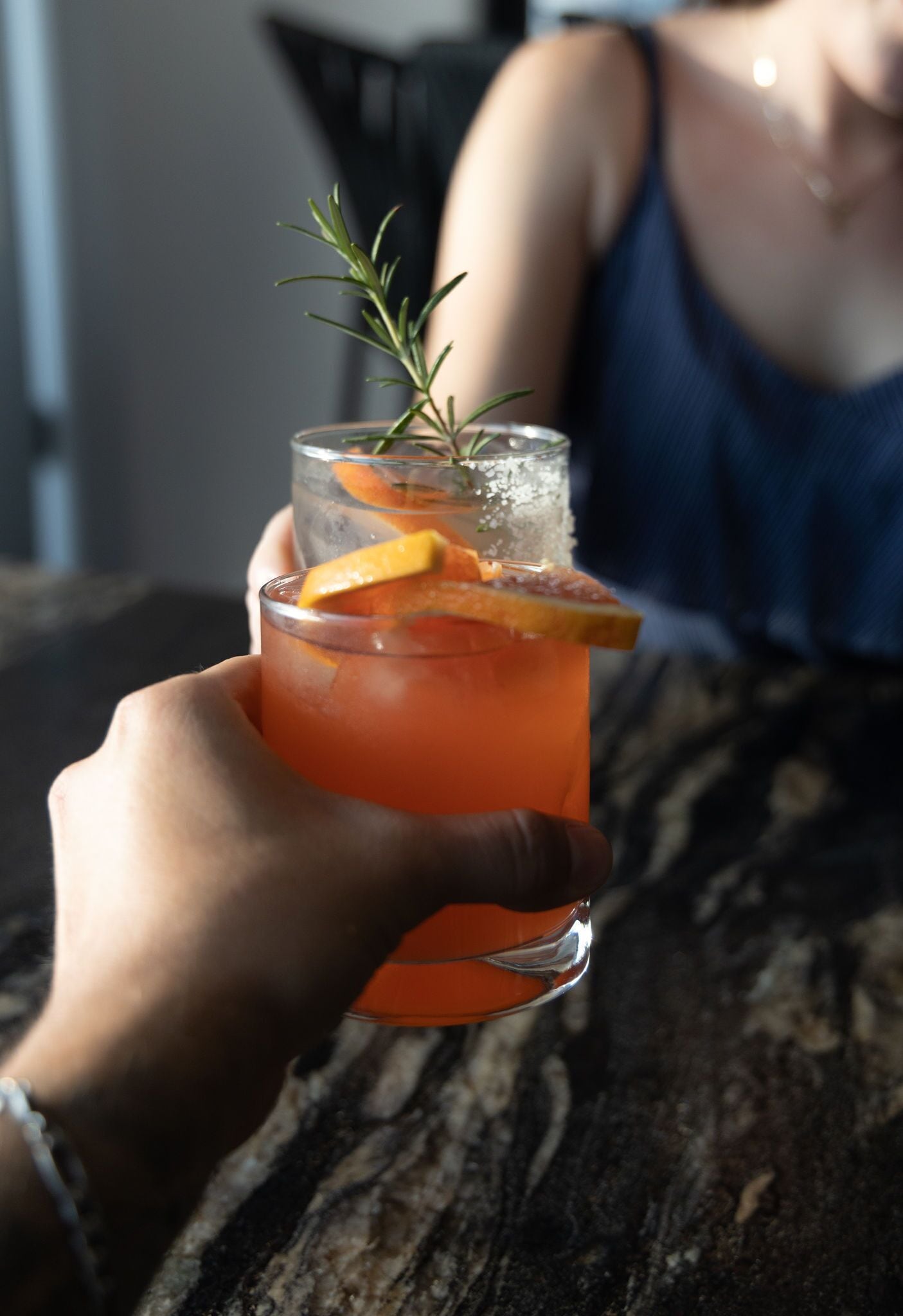 A hand holding a glass with a drink garnished with a slice of orange and a sprig of rosemary.