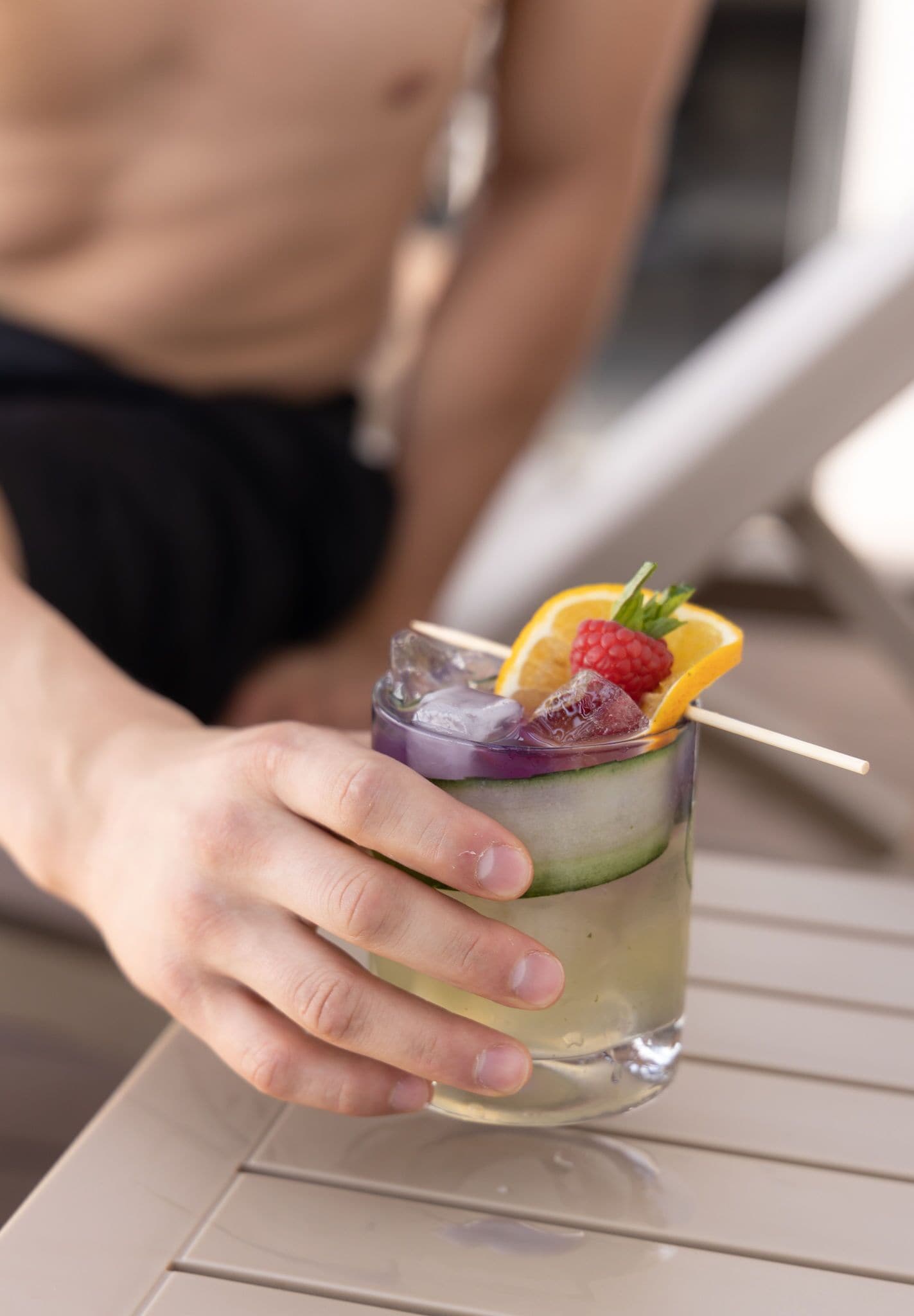 A person holding a cocktail with a cucumber slice, ice, and fruit garnish.