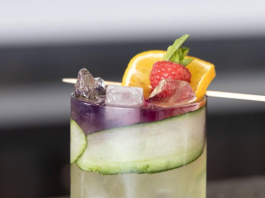 Een helder glas met een gelaagde drank, ijsblokjes, een aardbei en een stukje sinaasappel.