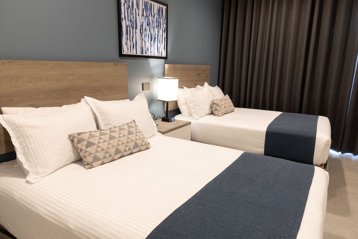 Two beds with white linens and a dark blue throw blanket, a nightstand with a lamp, and a framed abstract painting on the wall.