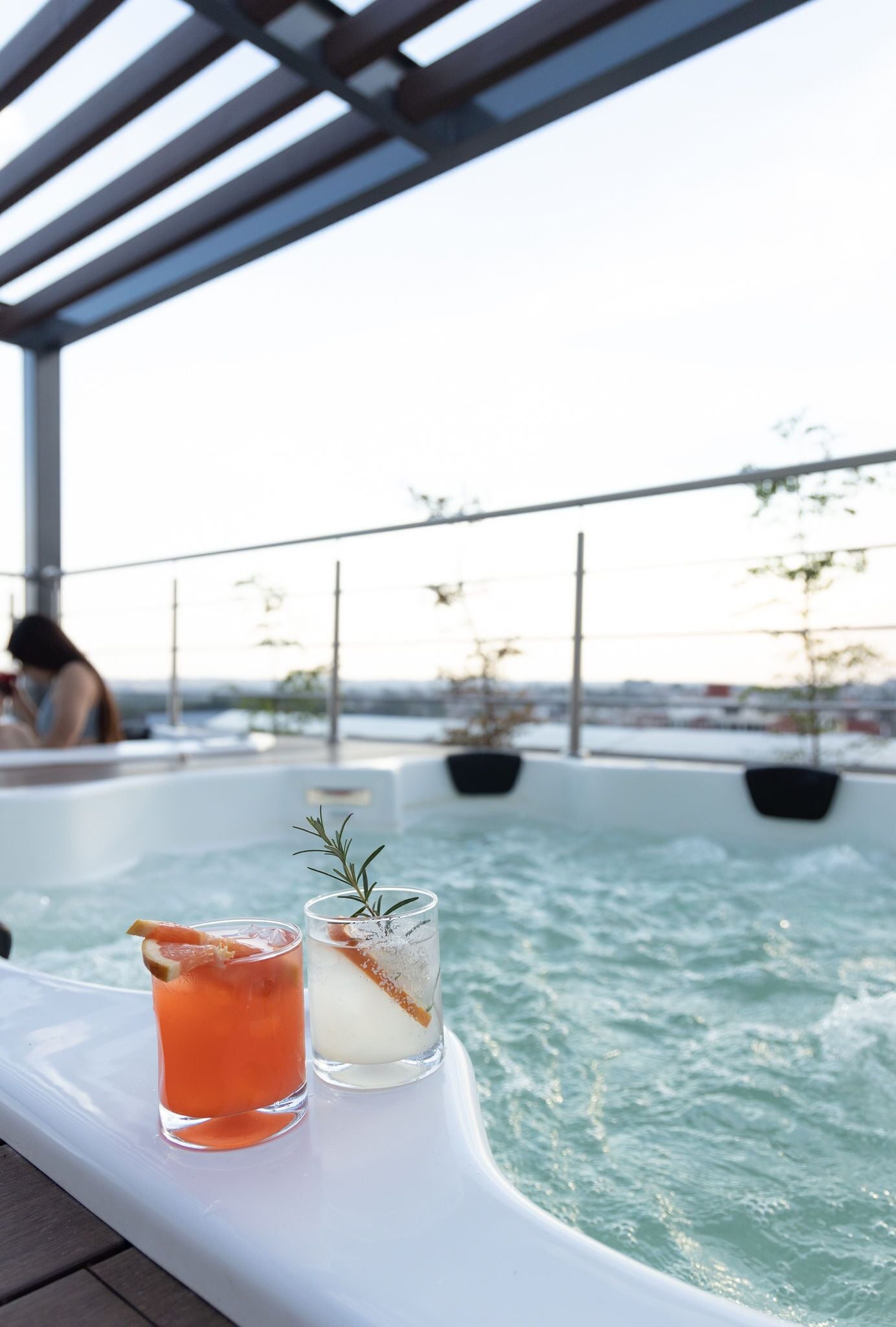 Zona de piscina con dos bebidas sobre una mesa blanca, una bañera de hidromasaje y vistas de la ciudad.